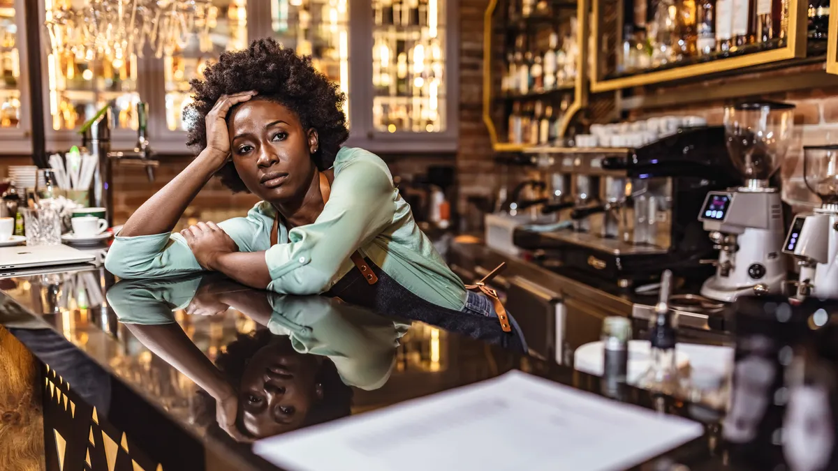 A female worker leans against the counter looking frustrated at a coffee bar with her head on her hand.