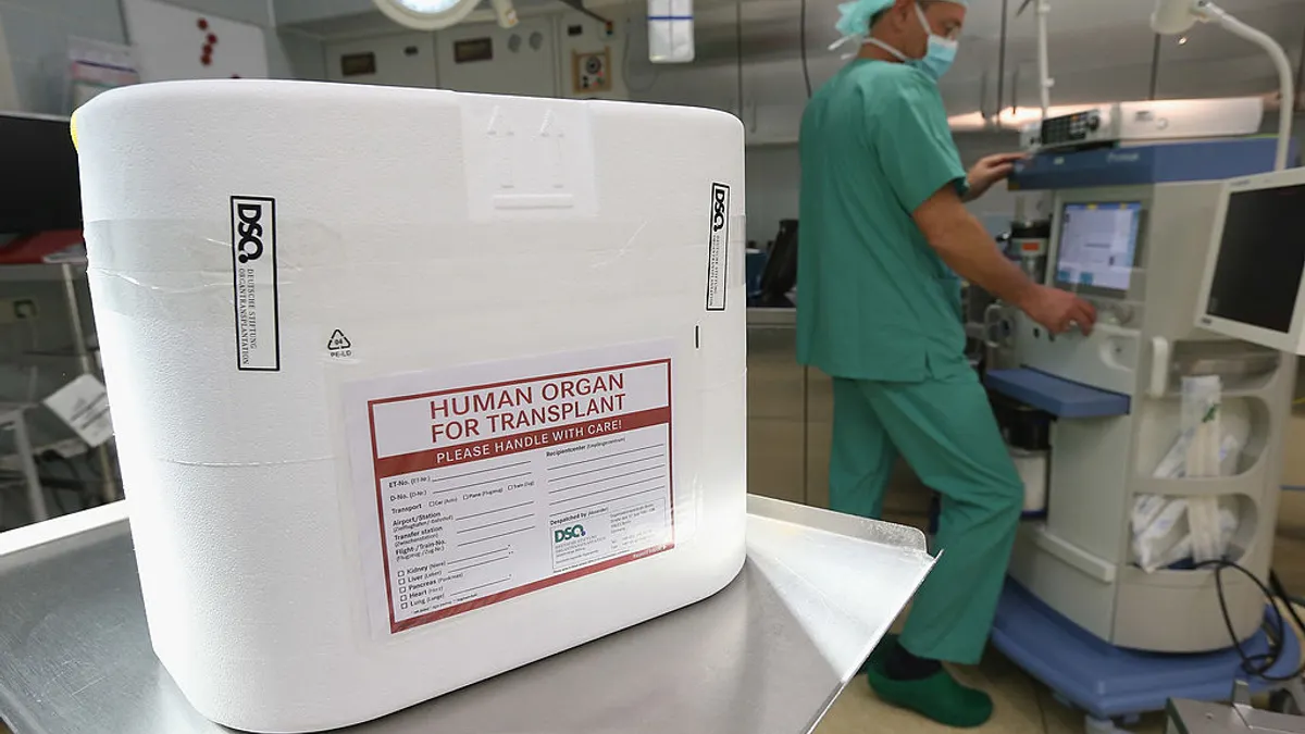 A cooler for an organ transplant sits on a cart in an operating room, as a hospital staffer works behind.
