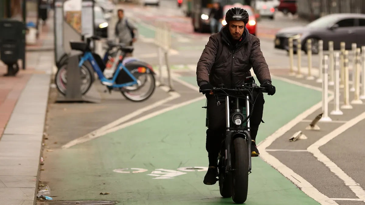 Man in heavy jacket with helmet rides an electric bike toward the camera on a green bike path.