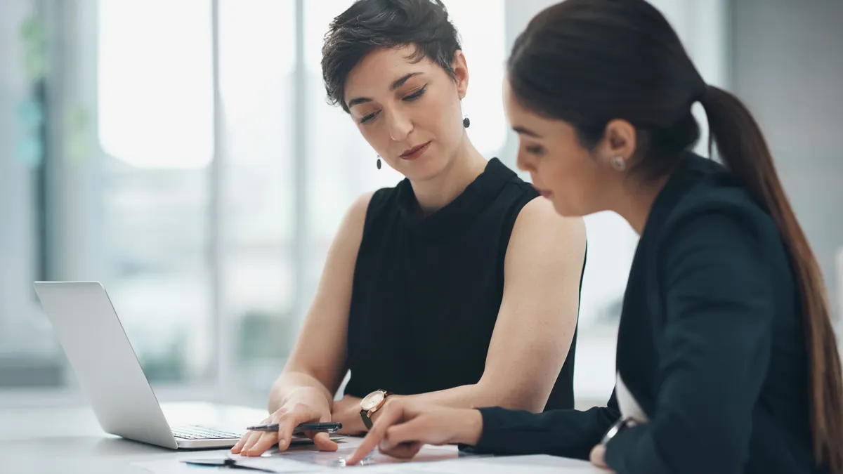 Two business people sitting at a desk looking at a document with a laptop on the table.