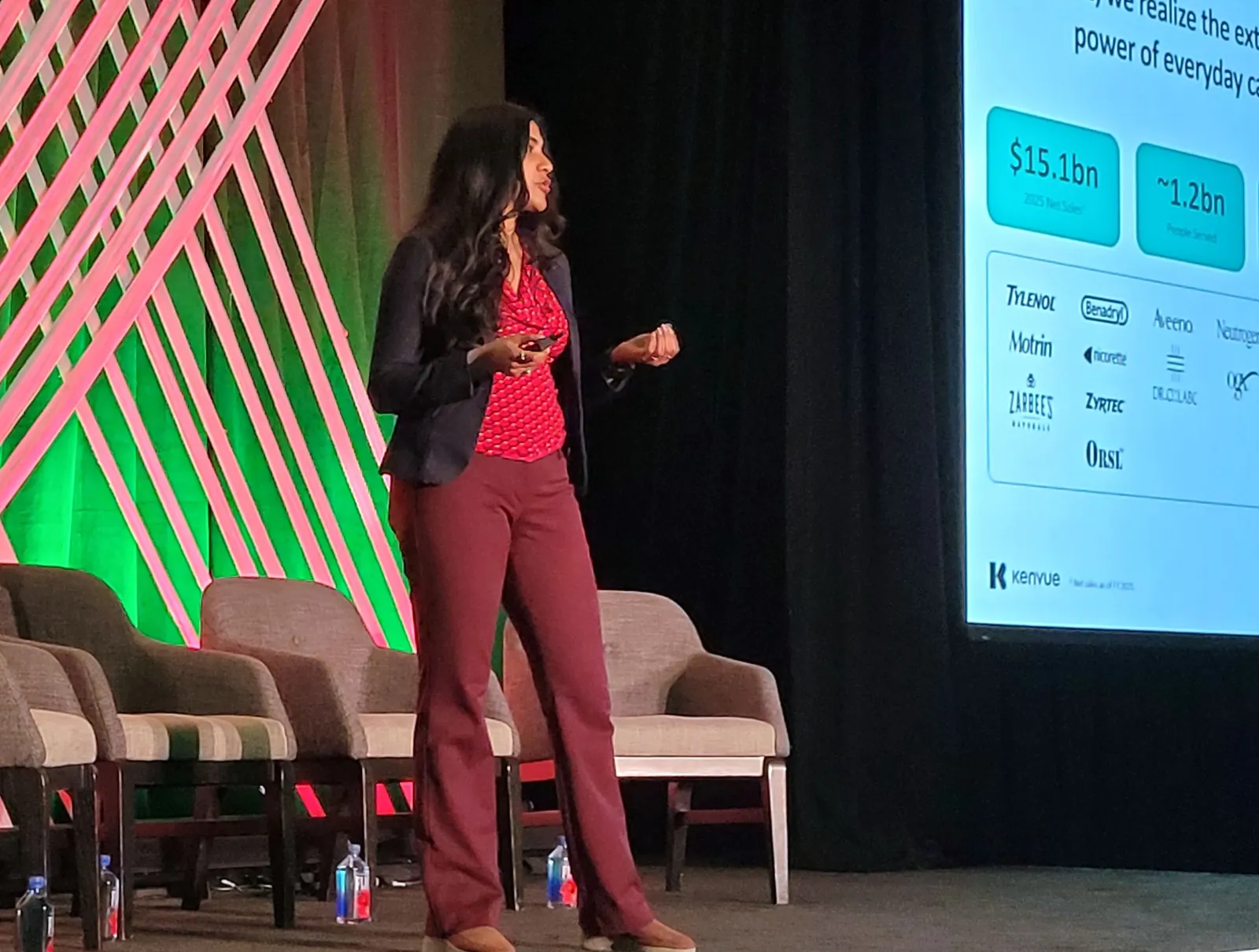 A person stands on a stage with a digital screen next to them while addressing an audience.