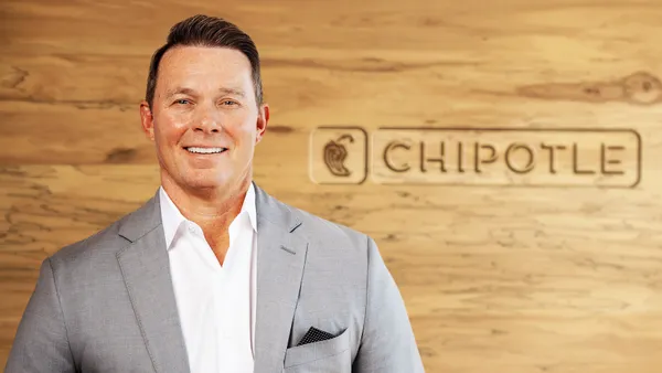 A man in a gray suit stands in front of a wood panel background with the logo "Chipotle."