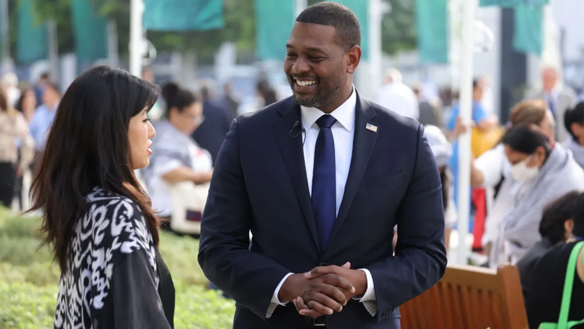 A woman speaks to a man wearing a suit with a U.S. flag pin outdoors, with flags behind them.