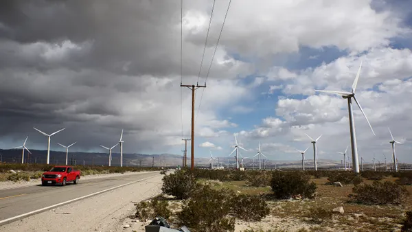Truck driving along road with a field of wind turbines stretching across the land behind it and a storm coming in from the left side of the sky.