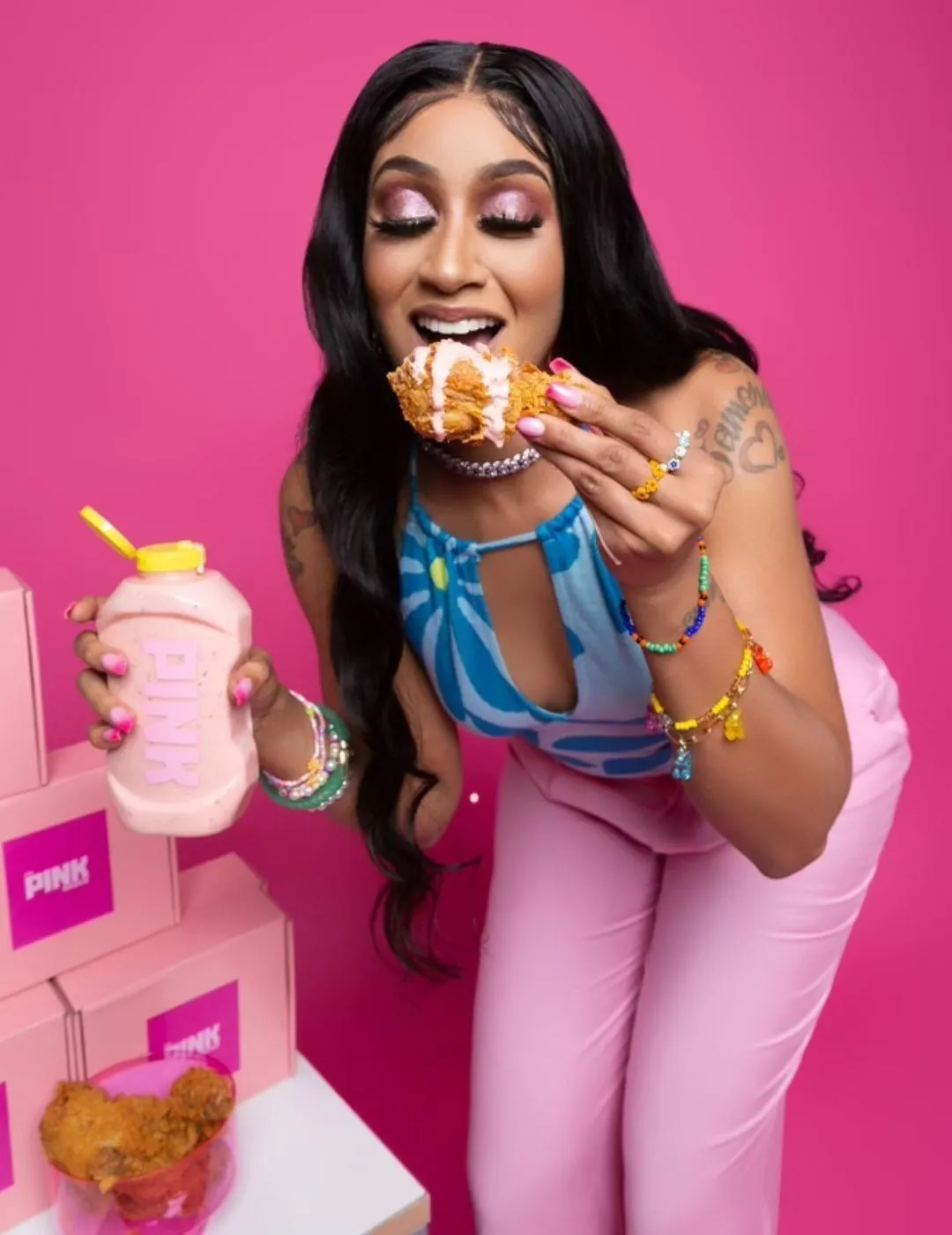 Pink Sauce creator Chef Pii bends at the waist, taking a bite of a fried chicken leg with Pink Sauce on top and holding a bottle of the condiment in her other hand.