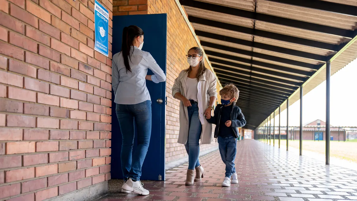 Teacher welcoming a student with her mother back to school and wearing facemasks