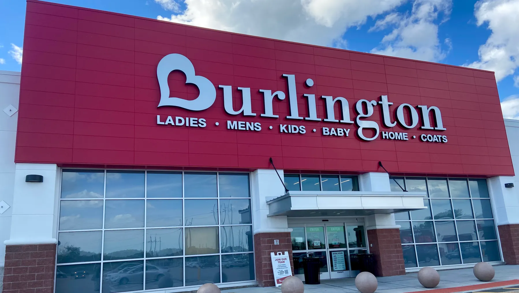 A red Burlington storefront says "ladies, mens, kids, baby, home, coats," as clouds billow overhead in a bright blue sky.