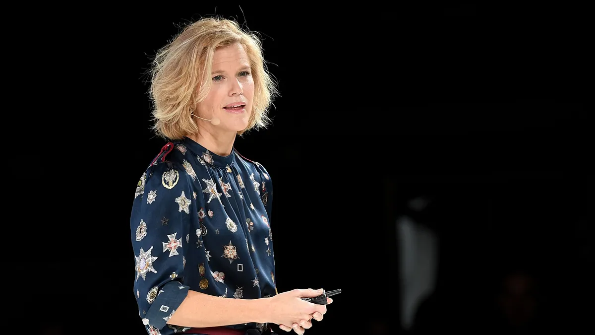 A woman in a blue jumpsuit printed with white medallions stands in front of a black screen, and she is turned slightly towards the camera