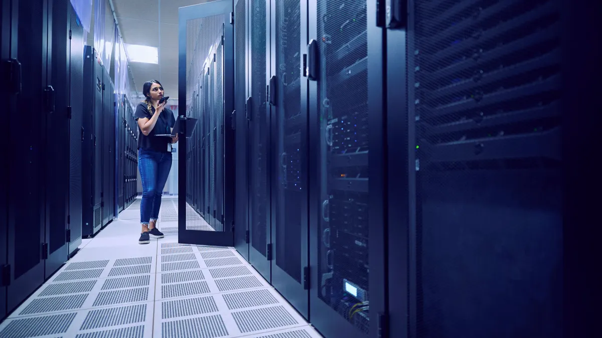 A person standing in front of a black server, holding a laptop on her left arm and speaking into her phone that's in her right hand.
