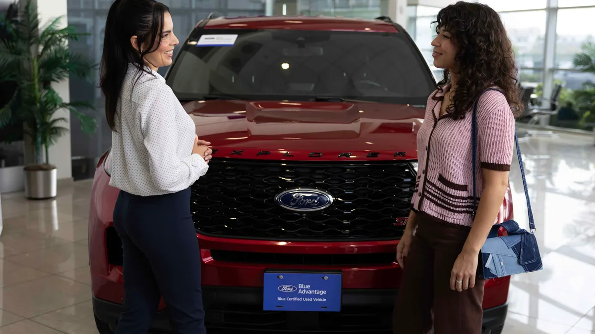 Two women standing in front of a red Explorer SUV in a Ford dealership showroom.