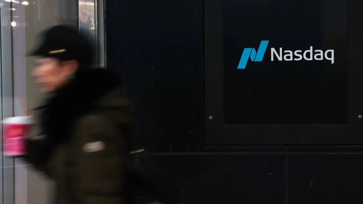 A sign spelling Nasdaq hangs on the side of a building while a pedestrian passes by.