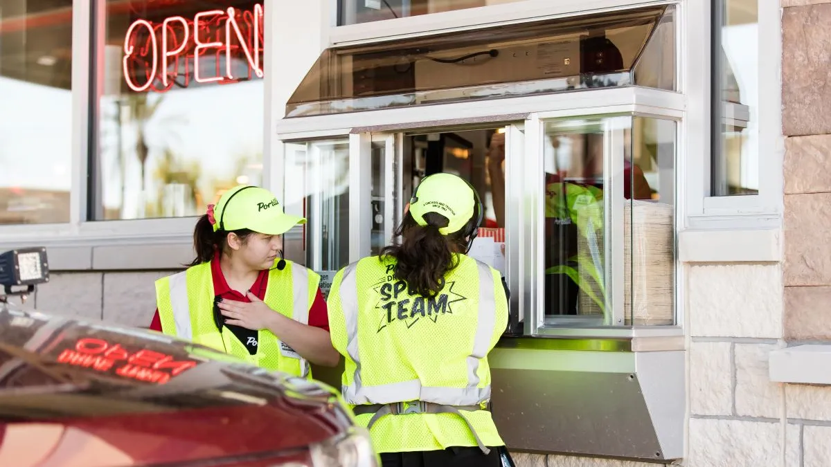 Two female Portillo's employees wearing yellow vests and serving customers at the drive-thru