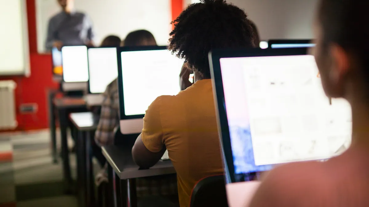 Students work on computers while a teacher instructs in front of them in a classroom.