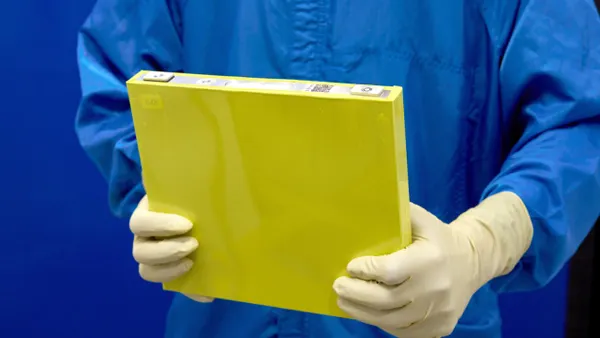 An employee wearing a blue protective suit holds a prototype LMR battery cell with a yellow housing at the General Motors Wallace Battery Cell Innovation Center in Warren, Michigan.