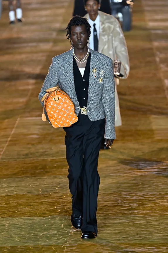 A man in a cropped gray jacket walks down a fashion runway holding an orange bag under his arm.