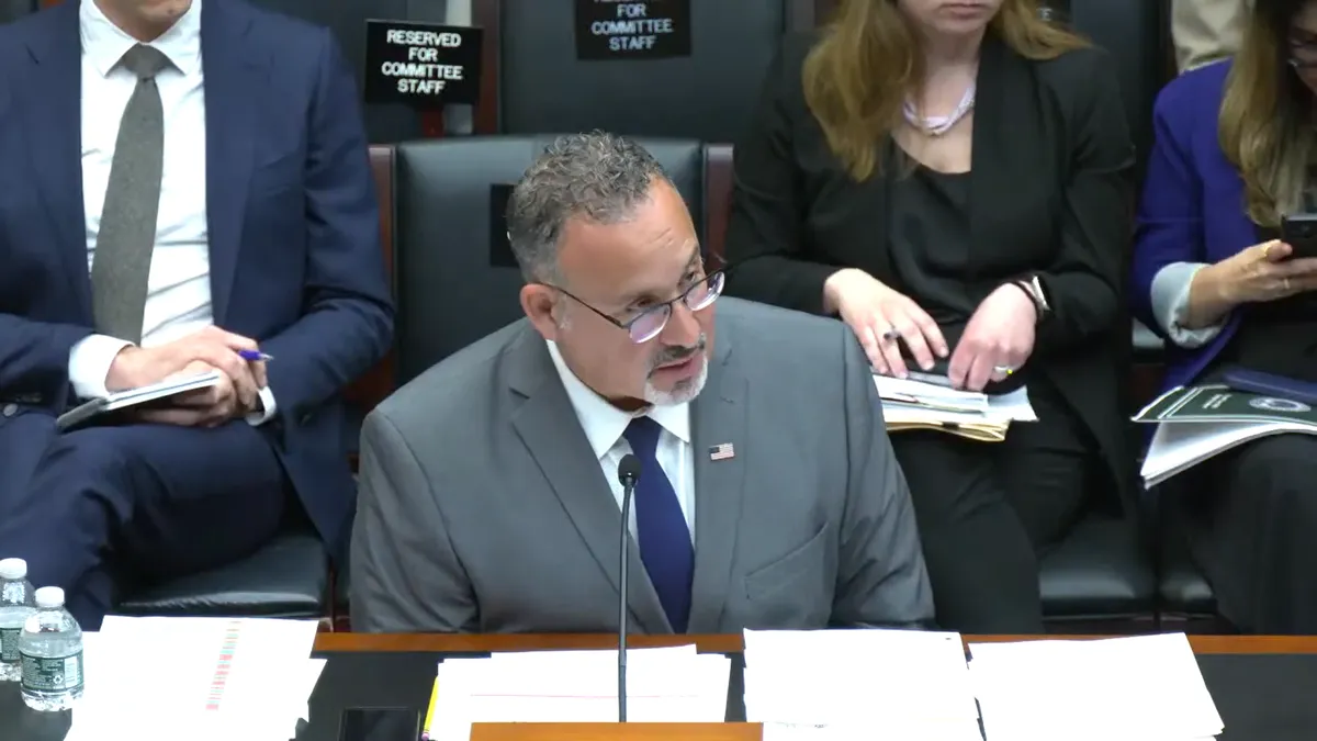 Education Secretary Miguel Cardona sits in a House hearing and speaks into a microphone