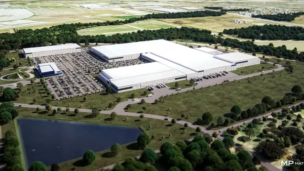 An area with a large white building next to a parking lot with cars, trees and grass.