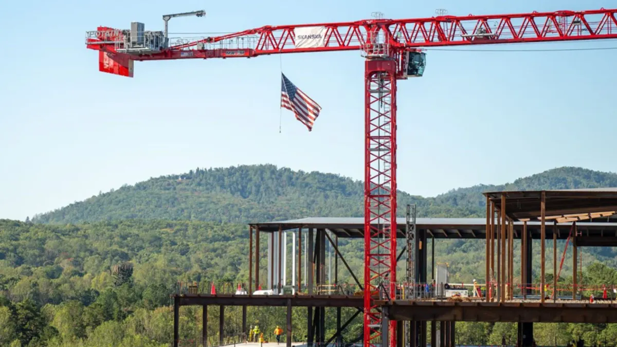 A crane looms above a steel structure of a building. In the background are large, verdant trees.