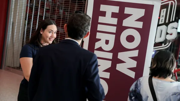Two people talk in front of a "now hiring" sign.