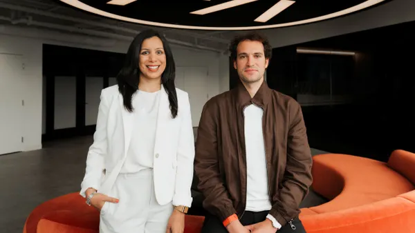 Two people are side by side. The person on the left wears a white suit and stands while the person on the right sits on the back of a couch.