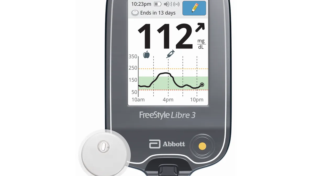 A grey reader shows a blood glucose reading, next to a white circular device