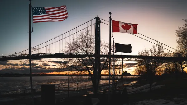 The Ambassador Bridge is shown with sunlight on the horizon and American and Canadian flags in the foreground.