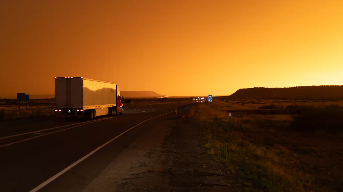 A tractor-trailer heads toward a sunrise with other traffic lights in the distance.