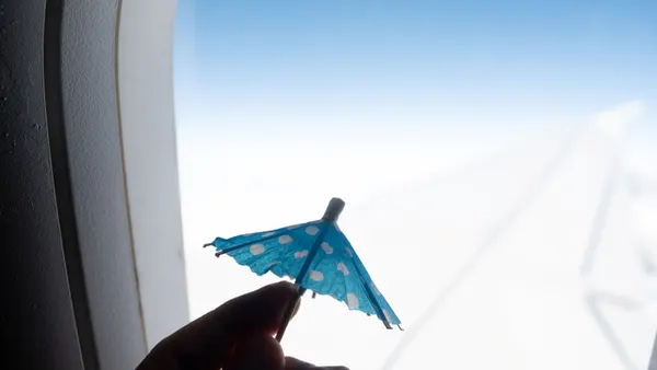 Man's hand holds a miniature beach umbrella in front of the window of an aircraft in flight.