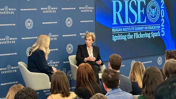 Two people are seated in chairs on a stage, angled toward each other. There are people sitting in chairs in rows in front on them. A backdrop and screen behind the speakers says "RISE"