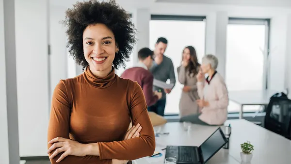A person smiling with a group huddle in the background.