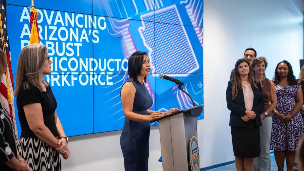 A person in a sleeveless blue dress speaking into a microphone in the center with people on their left and right.