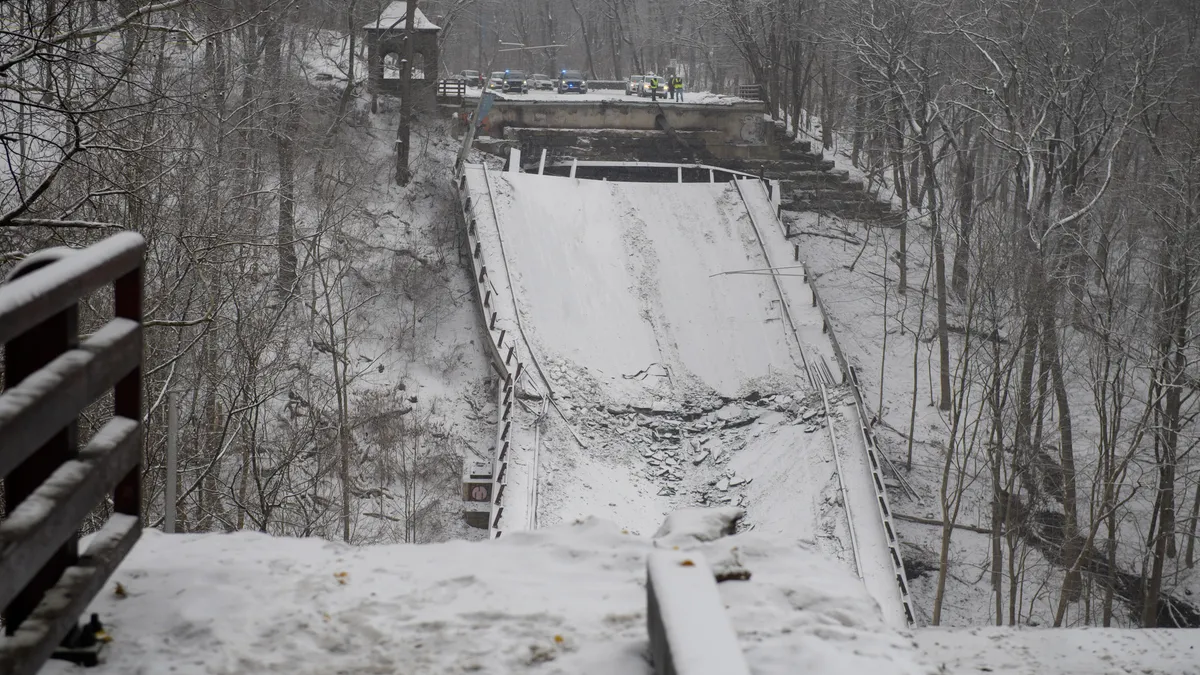 The broken edge of a snow-covered road bridge in the foreground with other bridge pieces visible in the middle ground and vehicles on an elevated structure in the distance.