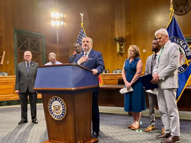 A person stands at a podium with a speaker in a wood-paneled room. A handful of other people are standing behind the person.
