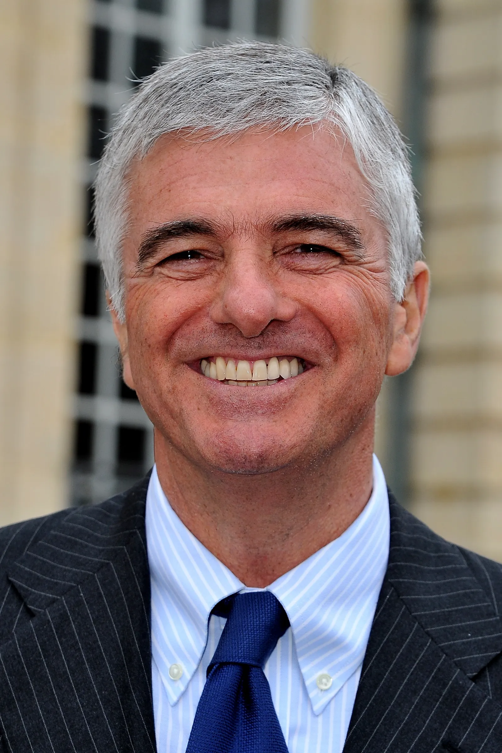 A person with short white hair wears a suit and grins widely at the camera.