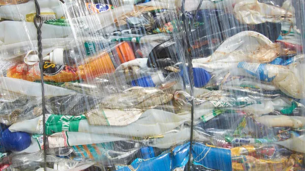 A tightly packed bale of crushed plastic bottles, compressed into a rectangular block and wrapped in clear plastic film. The bale contains various colored plastic bottles, flattened and compacted together, ready for recycling.