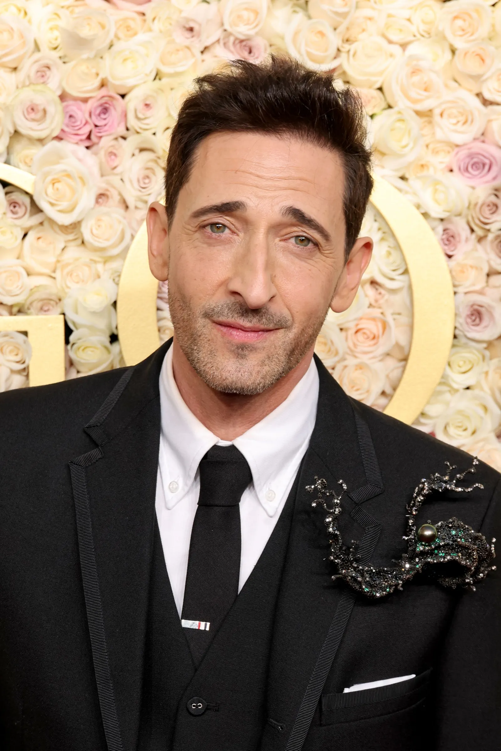 Person stares at the camera while wearing a black suit, white shirt, and large bejeweled lapel pin.