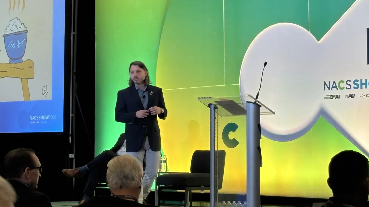 A person on stage speaks to a growd. Text on the screen behind them says  NACS Show.