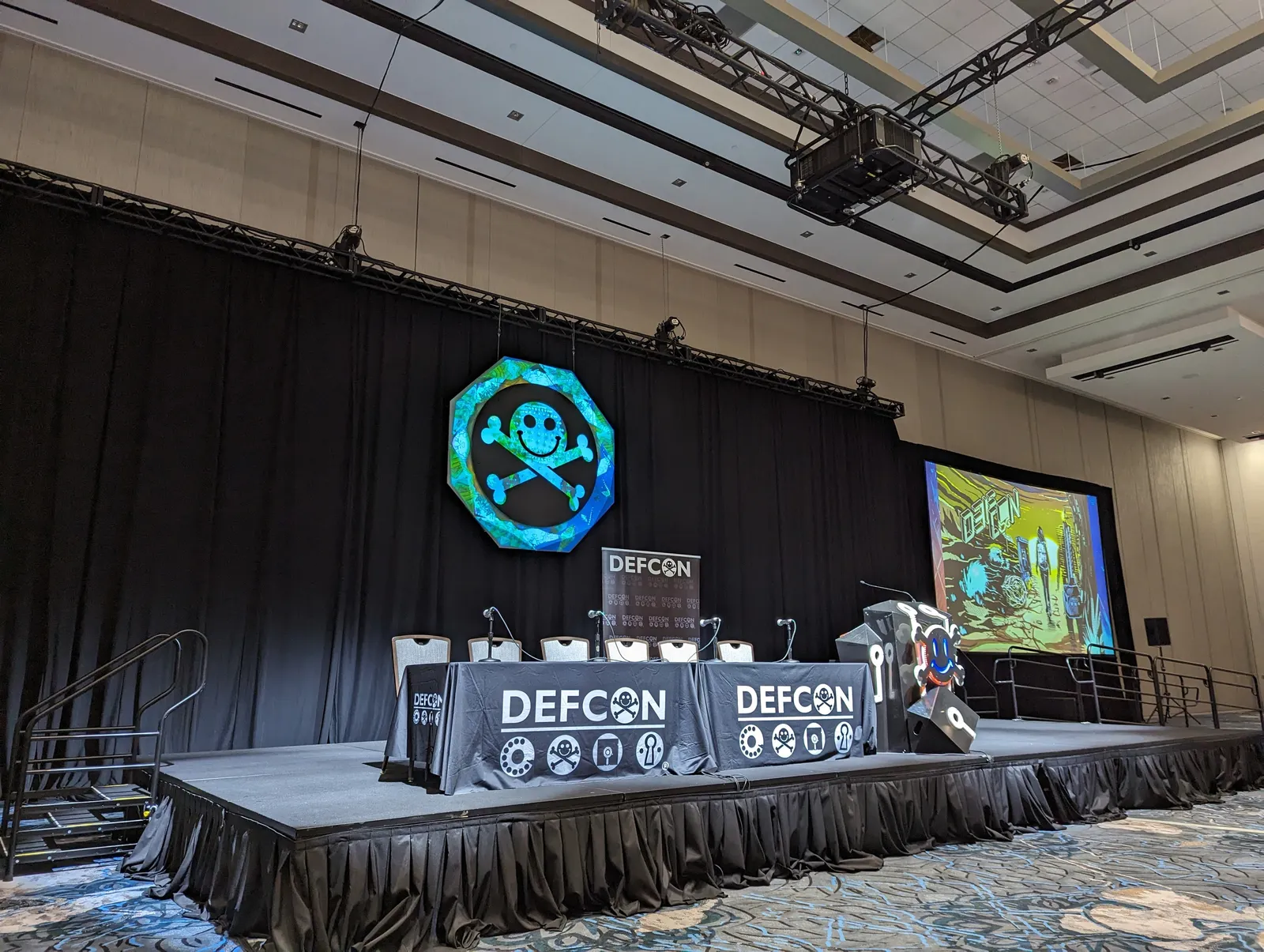 A table bearing DEF CON signage sits on a raised platform. Six chairs and a lectern also sit on the platform. The lectern is adorned with DEF CON-related paraphernalia. DEF CON's skull-and-crossbones logo hangs in front of a black curtain at the back of the room. To the right of the raised platform, a screen shows DEF CON-related artwork.