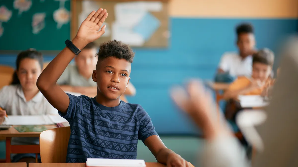 A Black boy seated at a desk raises his hand in a middle school classroom.