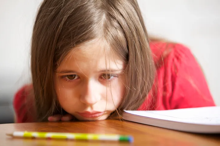 A child has their chin on a desk and looks sad. In front of the child on the desk is a pencil.