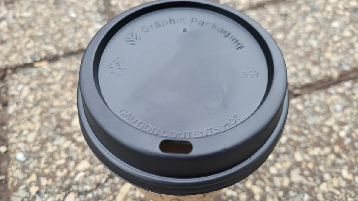 A close-up view of a black hot cup lid with Graphic Packaging International's name and a symbol indicating polystyrene.