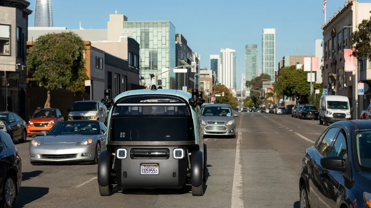 A blue vehicle with a large front window and sensors mounted on the upper sides seen on a busy urban street.
