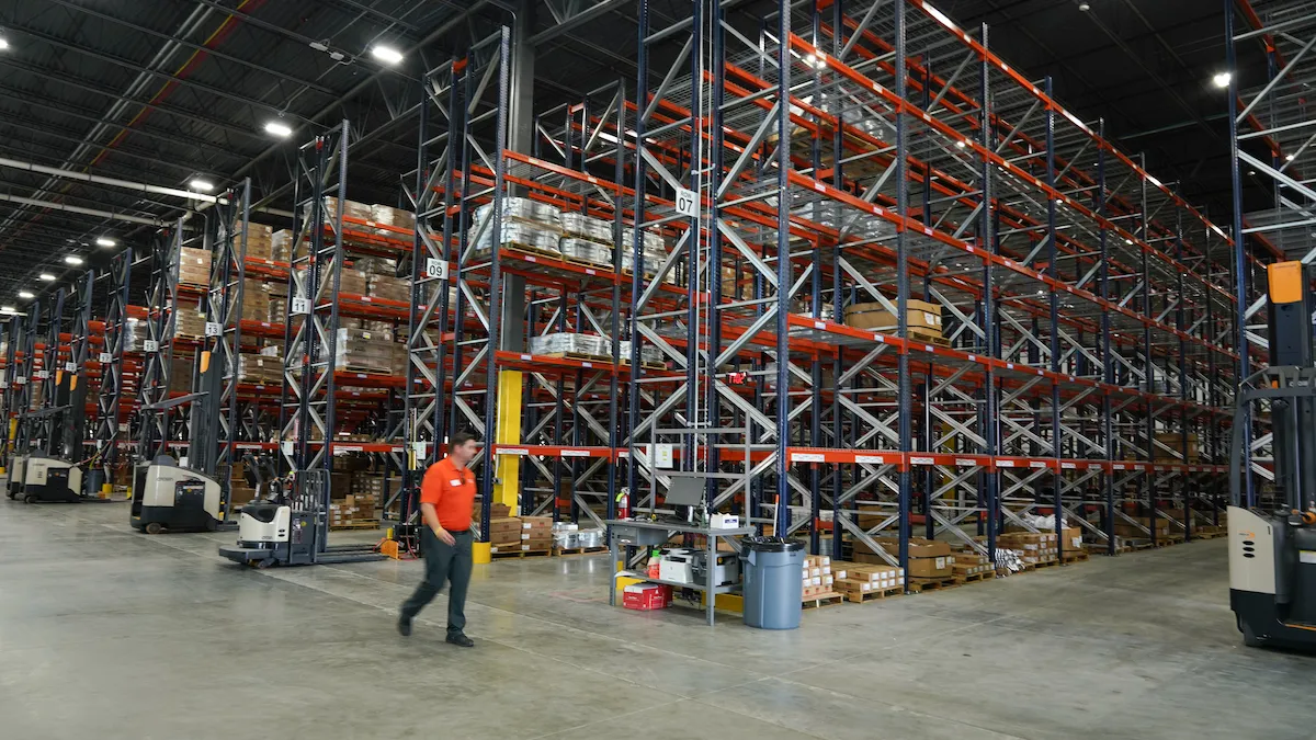 A man in an orange shirt walks around an industrial warehouse. There is red scaffolding around him, that holds various materials.