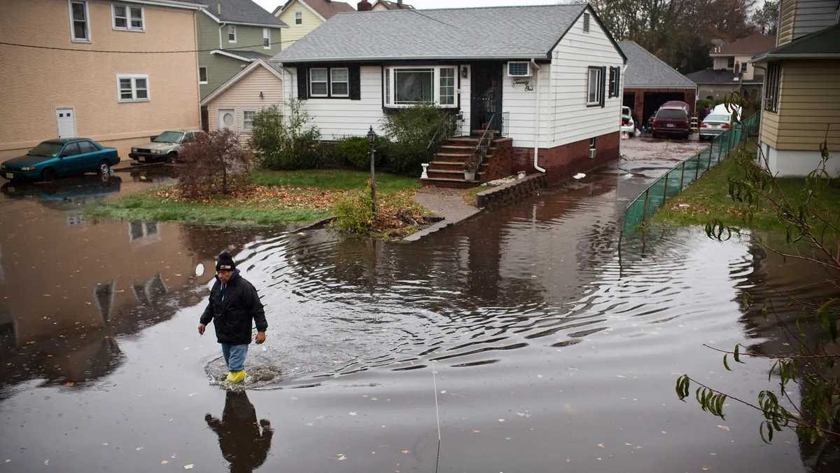 A person walks through knee-high floodwaters in front of a house.