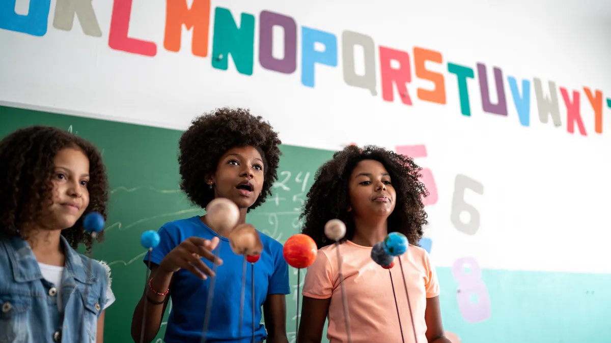 Three Black children deliver a science presentation in a classroom using a model of the solar system. A chalkboard with the letters of the alphabet above it is visible behind them.