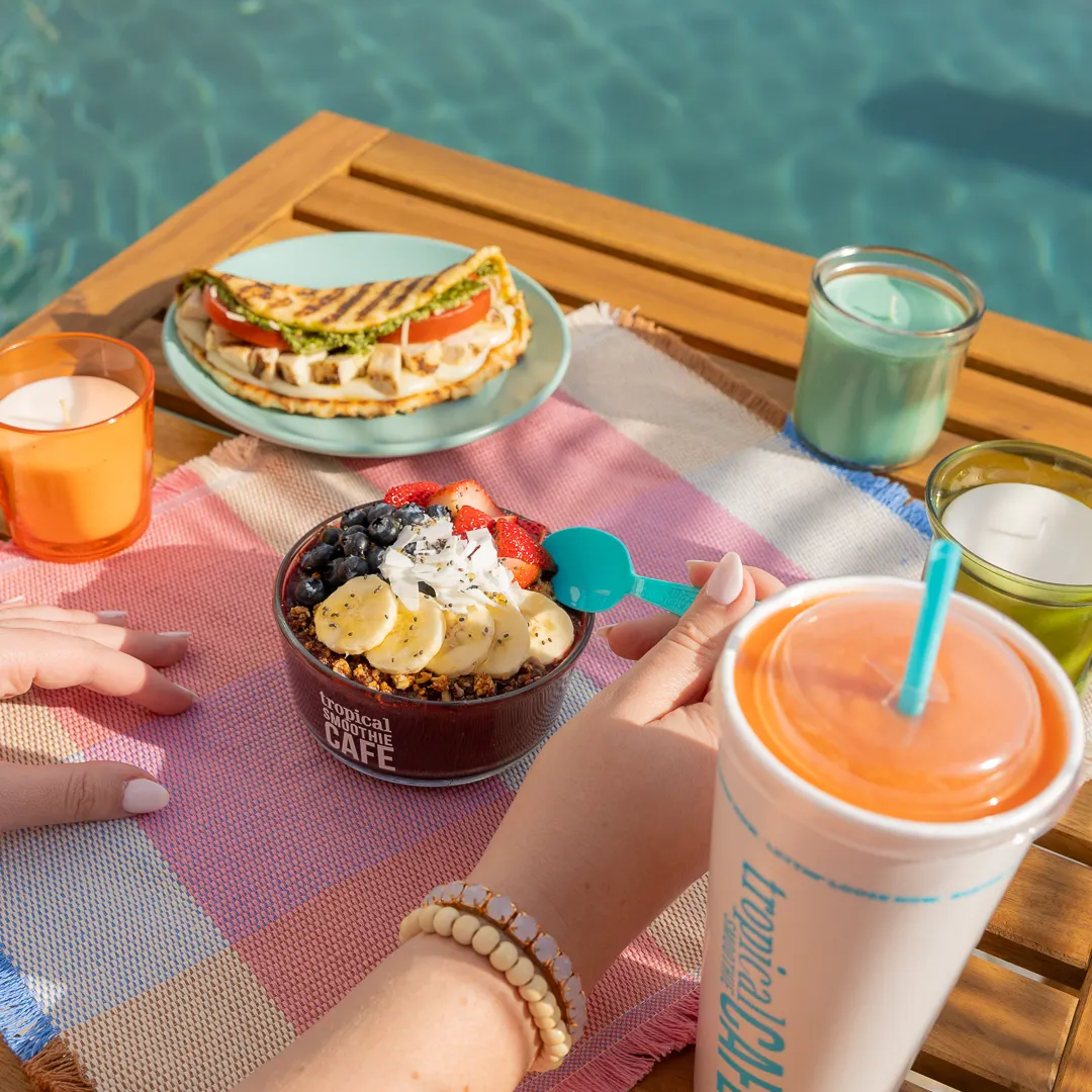 A photograph of a hand scooping a blue sppon into a bowl with bananans and berries, a blue plate with a flatbread and a white cup with an orange drink.
