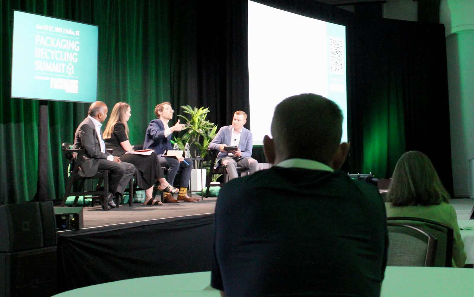 Four speakers sit in chairs on a stage in front of a background that says Packaging Recycling Summit while audience members sit at tables.