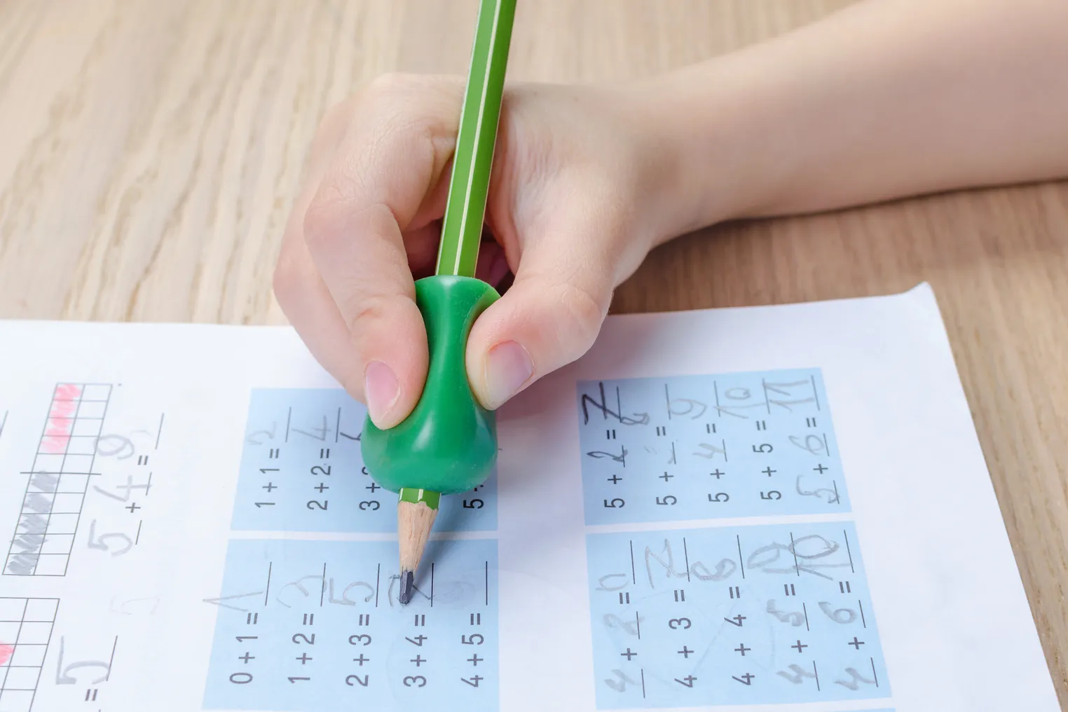 A close-up of a student holding a pencil with a finger grip. The pencil is pointed at a paper on a desk.