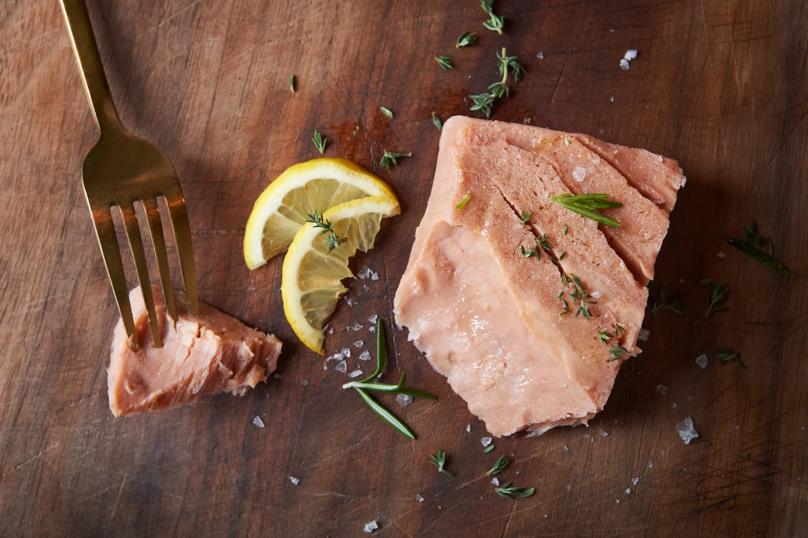 A fork has a piece of a New School Foods salmon filet on top of a wooden cutting board. Lemon slices and herbs and the rest of the salmon filet are next to it.