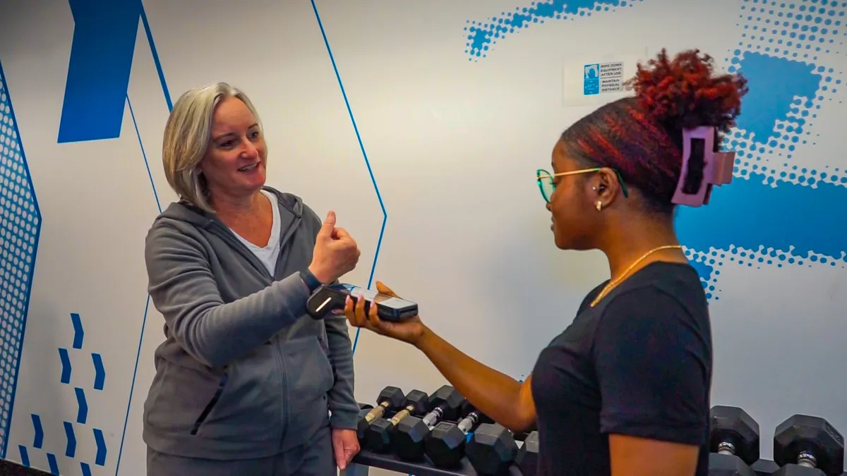 One person holds out a hardware device and the other person taps a watch to the device, with fitness center weights in the background.